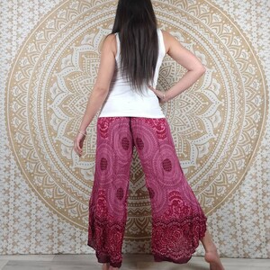 Puede incluir: Una persona con una camiseta sin mangas blanca y pantalones anchos magenta con un patr&oacute;n circular. Los pantalones tienen un borde magenta m&aacute;s oscuro. El fondo es un tapiz de mandala dorado.