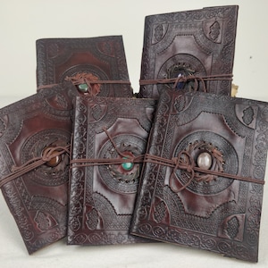 Op de afbeelding: Vijf bruine lederen dagboeken met decoratieve gegraveerde ontwerpen en edelsteenaccenten. Elk dagboek is dichtgebonden met een bruin koord.