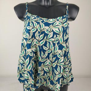 Puede incluir: Una blusa sin mangas con tirantes finos, con un fondo azul y un estampado repetido de hojas blancas, verdes y beige. La blusa tiene una silueta acampanada.