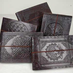 May include: Several dark brown leather journals with embossed designs. The journals display Celtic knots, a tree of life, and geometric patterns. Each journal is secured with a brown elastic band. The journals are made of leather.