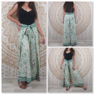 Op de afbeelding: Een wijde broek met een bloemenprint in tinten groen en wit. De broek heeft een wikkel-stijl en wordt in de taille vastgebonden met een sjerp. De broek wordt getoond op een model met een zwart topje.