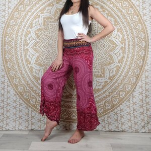 Puede incluir: Pantalones anchos magenta con un estampado de mandala y una cintura estampada. Los pantalones tienen un dobladillo acampanado y se combinan con una camiseta sin mangas blanca. El fondo presenta un tapiz de mandala dorado y blanco.