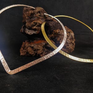 May include: Two open-ended, leaf-shaped earrings. One is silver-toned with a hammered texture, and the other is gold-toned. The earrings are displayed against a dark background with a piece of textured wood.