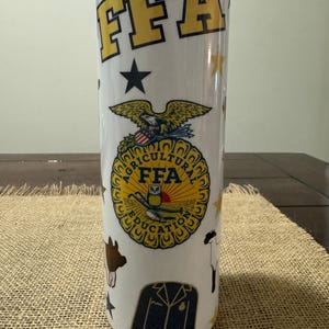 Op de afbeelding: Een witte roestvrijstalen tumbler met een geel en zwart FFA-logo, een zwart en gouden jack en andere landbouwbeelden. De tumbler heeft een doorzichtig deksel.