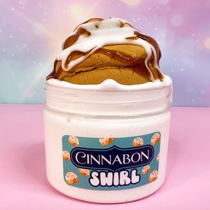 May include: A jar of white slime topped with a cinnamon roll, drizzled with white icing and caramel sauce. The jar has a label that reads "Cinnabon Swirl" with cinnamon roll graphics. The background is a gradient of pink, blue, and white.