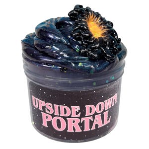 May include: A clear plastic container filled with dark blue slime, speckled with glitter, and topped with a black and orange swirl. The container has the words "UPSIDE DOWN PORTAL" in pink letters on a black background.