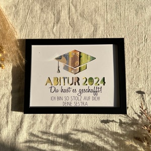 May include: A black framed picture with a white background. The picture has a graduation cap cut out of it, with money tucked inside. The text "ABITUR 2024" is at the top of the picture, followed by "Du hast es geschafft!" and "ICH BIN SO STOLZ AUF DICH! DEINE SESTRA".