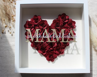 Puede incluir: Un marco blanco con un corazón de flores de papel rojo que dice "MAMA" en letras doradas con el texto "Ich habe dich lieb" en escritura cursiva blanca.