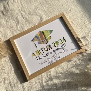 May include: A white framed picture with a graduation cap cutout. The text reads "ABITUR 2024 Du hast es geschafft! ICH BIN SO STOLZ AUF DICH! DEINE SESTRA"