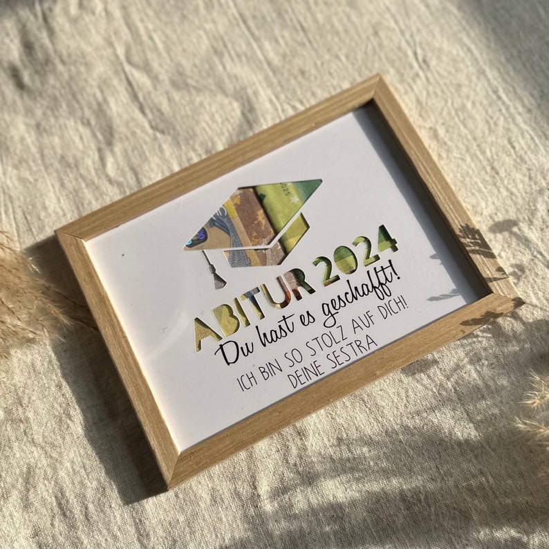 May include: A white framed picture with a graduation cap cutout. The text reads "ABITUR 2024 Du hast es geschafft! ICH BIN SO STOLZ AUF DICH! DEINE SESTRA"
