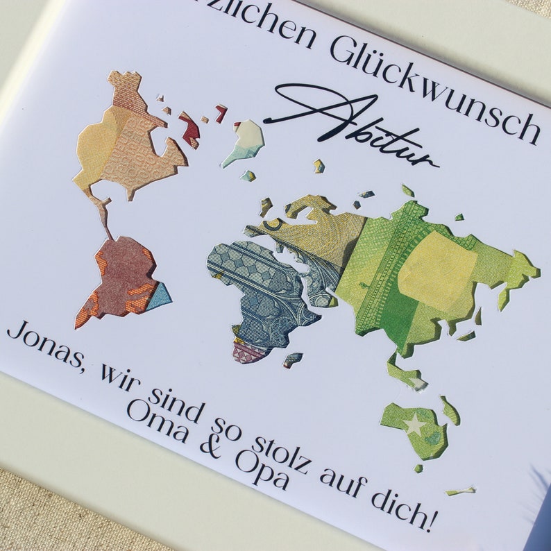 May include: A white card with a world map cut out of it. The map is made of different pieces of paper currency. The card has the text "lichen Gl&uuml;ckwunsch Abitur Jonas, wir sind so stolz auf dich! Oma & Opa" written on it.