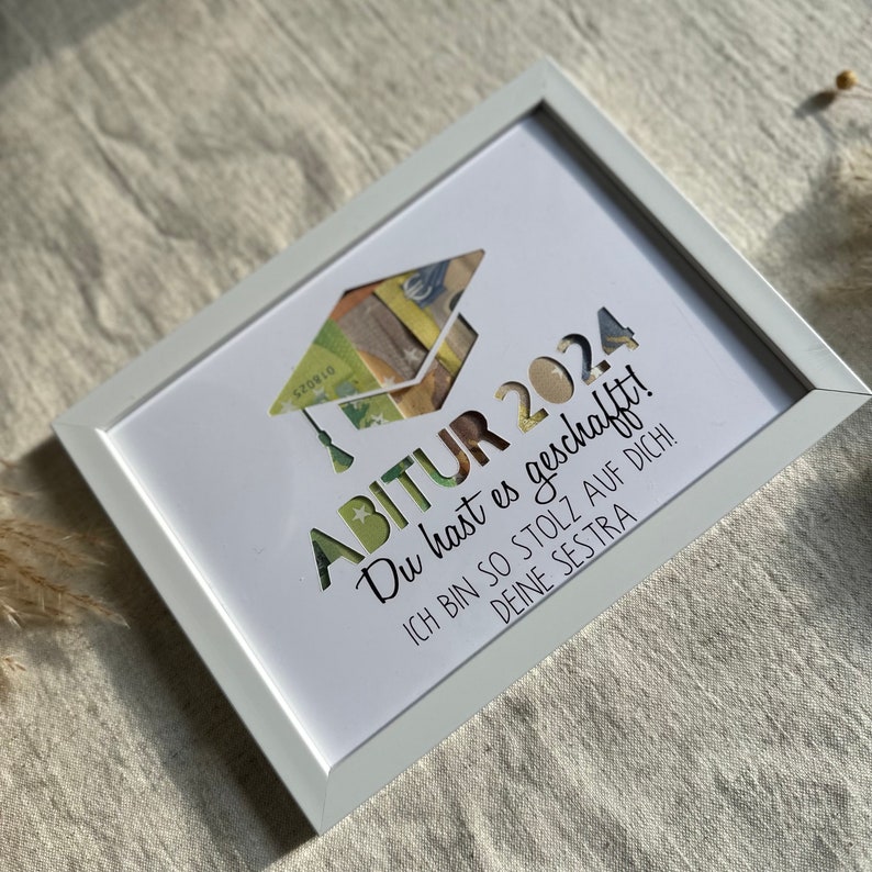 May include: A white picture frame with a white mat and a graduation cap made of money. The text in the image reads "ABITUR 2024 Du hast es geschafft! Ich bin so stolz auf dich! Deine Sestra"
