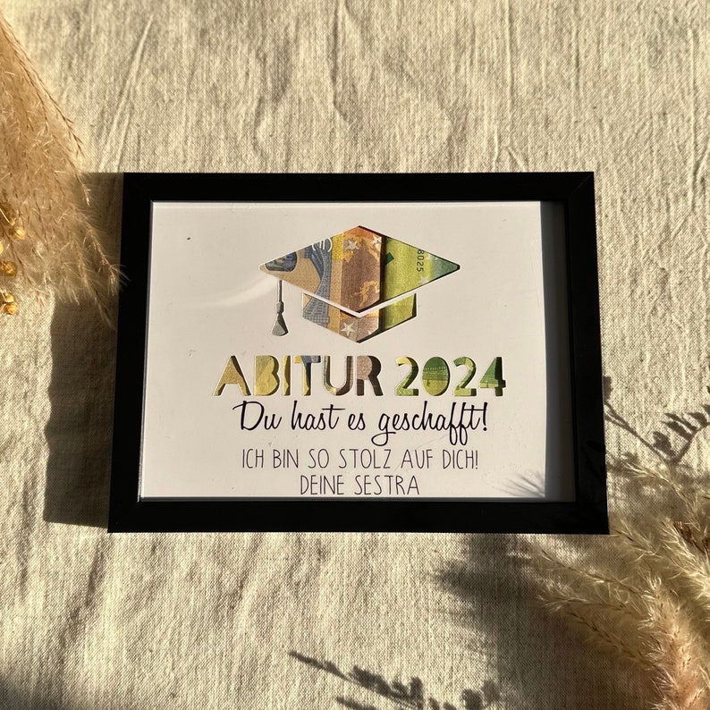 May include: A black framed picture with a white background. The picture has a graduation cap made of money with the text "ABITUR 2024" and the message "Du hast es geschafft! ICH BIN SO STOLZ AUF DICH! DEINE SESTRA".