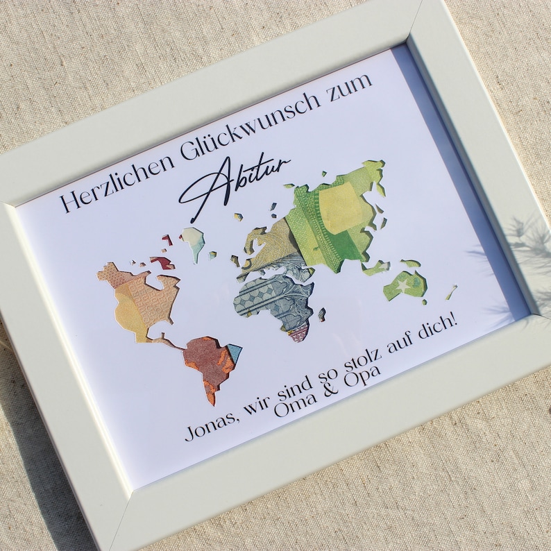 May include: A white framed picture with a world map made of money. The text "Herzlichen Gl&uuml;ckwunsch zum Abitur" is at the top of the picture. The text "Jonas, wir sind so stolz auf dich! Oma & Opa" is at the bottom of the picture.