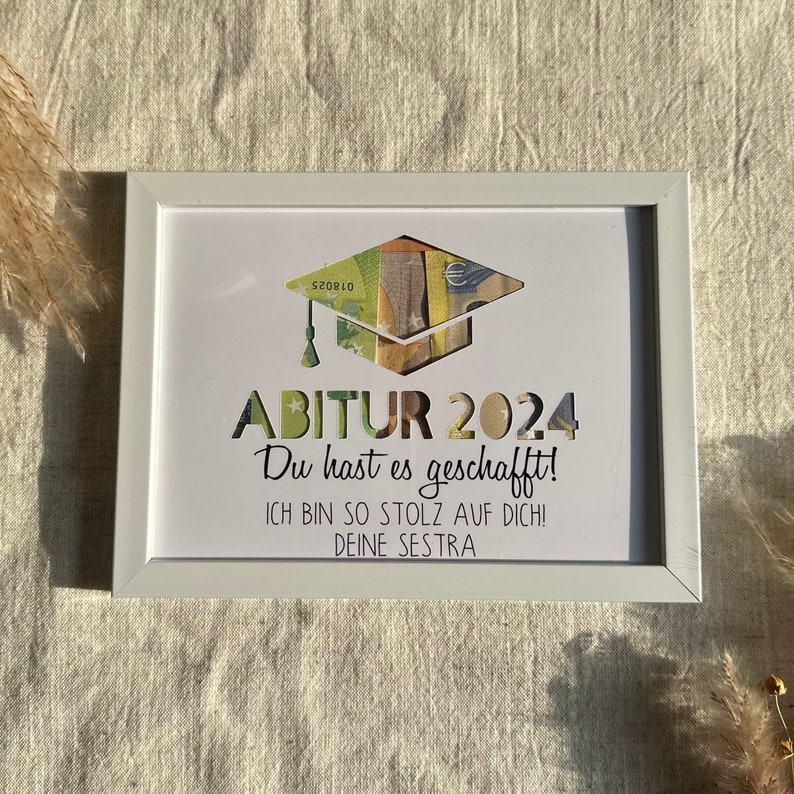 May include: A white picture frame with a white mat and a graduation cap made of money. The text in the frame reads "ABITUR 2024 Du hast es geschafft! ICH BIN SO STOLZ AUF DICH! DEINE SESTRA"