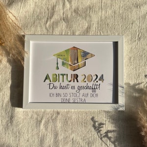 May include: A white picture frame with a graduation cap made of money inside. The text reads "ABITUR 2024 Du hast es geschafft! ICH BIN SO STOLZ AUF DICH! DEINE SESTRA"