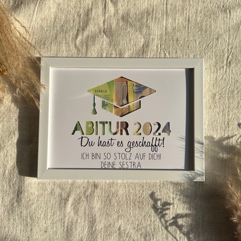 May include: A white picture frame with a graduation cap made of money inside. The text reads "ABITUR 2024 Du hast es geschafft! ICH BIN SO STOLZ AUF DICH! DEINE SESTRA"