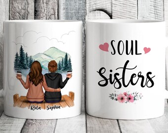 May include: Two white mugs with a design on each side. One mug features a watercolor illustration of two women sitting on a dock overlooking a lake and mountains. The text "Rula" and "Sophia" is printed below the women. The other mug features the text "SOUL SISTERS" with two pink hearts and a floral design.