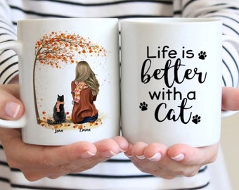 May include: White ceramic mug with a black cat and a woman sitting under a fall tree on one side and the text "Life is better with a cat" on the other side.
