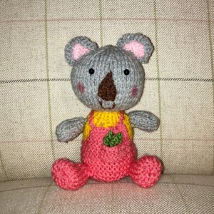 May include: A grey knitted koala wearing pink dungarees with a green leaf design. The koala has black eyes and a brown nose.