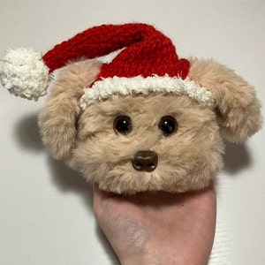 Puede incluir: Una cabeza de perro de peluche marrón claro con ojos grandes y oscuros y una nariz marrón. El perro lleva un gorro de Papá Noel rojo con ribete blanco y un pompón blanco esponjoso. El juguete es suave y peludo.