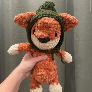 May include: A handmade orange and white crochet fox doll wearing a green hood. The fox has a white muzzle, a black nose, and white paws. The doll is soft and plush, with a textured appearance.
