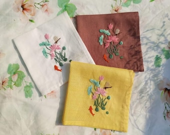 May include: Three embroidered linen tea towels with a floral design. The towels are white, brown, and yellow. Each towel features a pink lotus flower with green leaves and a dragonfly.