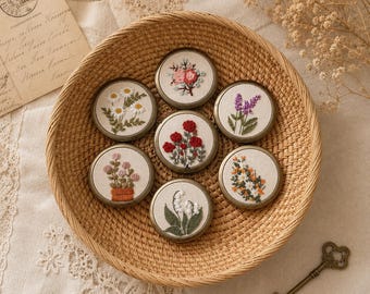 May include: A woven basket holds seven round, embroidered floral designs. Each design features a different flower, including daisies, roses, and lavender, in various colors. The embroidery is set against a cream-colored background with a gold-colored metal frame.