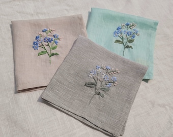 May include: Three embroidered linen handkerchiefs with a floral design. The handkerchiefs are in shades of pink, gray, and light blue.