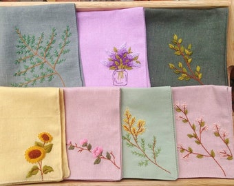 May include: Eight embroidered fabric squares with different floral designs. The squares are arranged in two rows of four. The designs include a sunflower, a sprig of flowers, a sprig of leaves, a bouquet of flowers in a vase, and a sprig of leaves.
