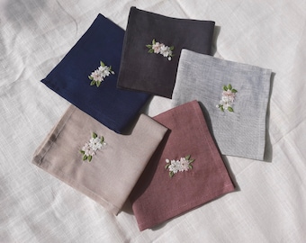 May include: Five embroidered linen napkins in different colors: navy blue, gray, beige, burgundy, and black. Each napkin features a delicate floral design with white flowers and green leaves.