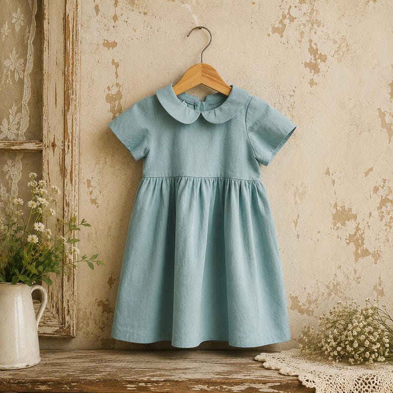 May include: A light blue short-sleeved dress featuring a rounded Peter Pan collar and a gathered waistline. The garment is displayed on a wooden hanger and has a simple, solid-colored design with a button closure visible at the back neckline.