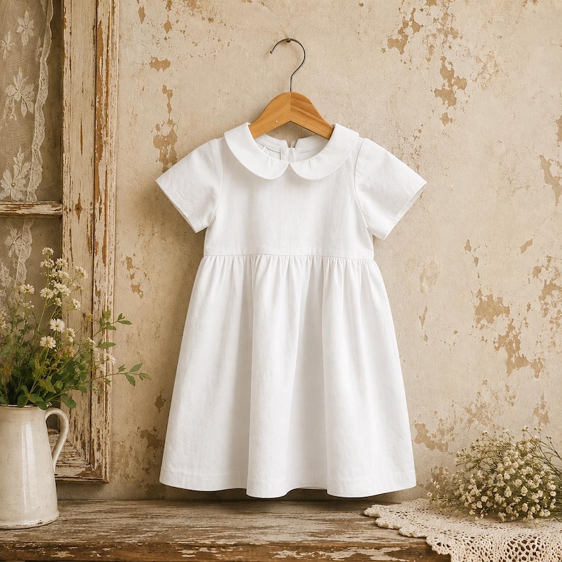 May include: A solid white short-sleeved dress featuring a rounded Peter Pan collar and a gathered waistline. The garment is displayed on a wooden hanger, showcasing a simple design with a flared skirt and a clean, plain white fabric finish.