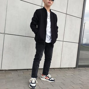 May include: A person wearing a black bomber jacket, white shirt, black jeans, and sunglasses. The person is also wearing sneakers with white, black, and red accents. The outfit is casual and stylish.