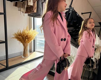 May include: A pink suit with a double-breasted blazer and wide-leg pants. The suit is worn with black shoes and a small black handbag. The outfit is displayed in a boutique setting with other clothing visible in the background.