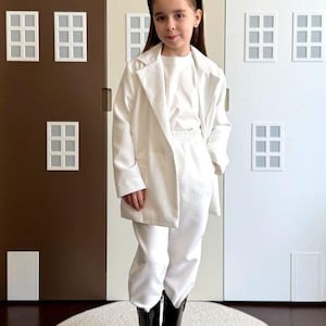 Puede incluir: Una persona con una blazer blanca, pantalones a juego y botas negras, de pie sobre una alfombra blanca. El fondo presenta una pared con paneles marrones, beige y blancos, decorada con recortes blancos en forma de ventana.