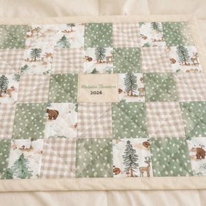 Op de afbeelding: Een patchwork quilt met een crèmekleurige rand. De quilt heeft vierkanten van groene stippen, beige ruitjes en stof met bosdieren- en boomontwerpen. Een label in het midden luidt "Madeline Theodore 2026".