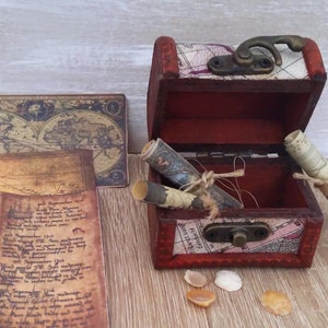 Cofre del tesoro pirata con 3 pergaminos: mapa del mundo antiguo, diseño de barco pirata y diario del capitán.