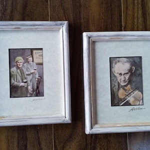 Könnte beinhalten: Zwei gerahmte Aquarellbilder. Das erste Bild zeigt zwei Männer in einem Pub, einer hält ein Bierglas. Das zweite Bild zeigt einen Mann, der Geige spielt. Beide Bilder sind mit "Ashe." signiert.