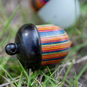 Op de afbeelding: Een klein, houten eikelvormig tolletje met een zwarte dop en een kleurrijk gestreept lichaam. De tol is gemaakt van hout en heeft een natuurlijke afwerking. De strepen zijn in verschillende kleuren, waaronder rood, oranje, geel, groen, blauw en paars.