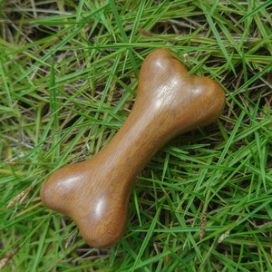 Op de afbeelding: Een houten hondenbot speelgoed, gesneden in de vorm van een bot, met een gladde, gepolijste afwerking. Het bot is lichtbruin van kleur en ligt op een bed van groen gras.