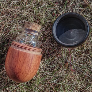 May include: A wooden container with a glass bottle inside. The bottle has a cork stopper and is sitting on a bed of dry grass. The container is a light brown color and has a smooth, rounded shape.