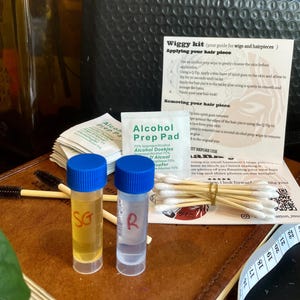 May include: A wig care kit featuring a brown glass bottle, two small vials with blue caps, and a black bubble mailer. The kit includes alcohol prep pads, cotton buds, and a guide for applying and removing hair pieces.