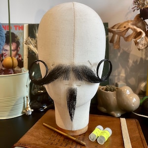 May include: A beige fabric-covered mannequin head displaying a black handlebar mustache and a small black goatee. The facial hair is pinned to the head, which rests on a brown leather-bound notebook alongside a gold-toned stylus and two small vials.