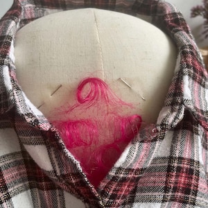 May include: A plaid button-down shirt featuring a pattern of white, black, and muted red lines. The shirt is draped over a beige fabric dress form, with two silver sewing pins securing the collar and a cluster of bright pink synthetic hair.