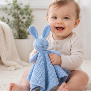 Peut inclure: Une couverture de sécurité en crochet bleu clair avec une tête de lapin en peluche aux longues oreilles et aux yeux fermés brodés. Le corps de la couverture présente un motif carré texturé, offrant une surface douce et tactile.