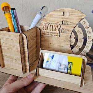 Peut inclure: Organisateur de bureau en bois découpé au laser avec un porte-stylo, un porte-cartes et un calendrier perpétuel. L'organisateur porte l'inscription "SUCCESS" et le texte "IN THE YEAR OF YOUR DREAMS". Comprend des stylos et une carte Metro.
