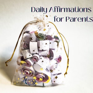 May include: A white mesh bag with a gold drawstring filled with rolled up pieces of paper with gold stars and moons printed on them. The bag is labeled "Daily Affirmations for Parents".