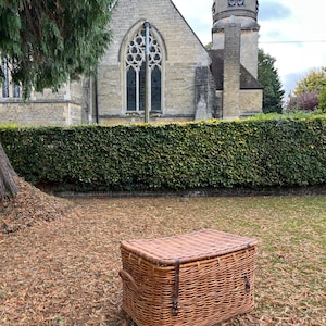 May include: A rectangular wicker trunk with a hinged lid and leather straps. The trunk is brown and rests on fallen leaves. A stone church with a tall arched window is in the background. The trunk is approximately 76 cm long.