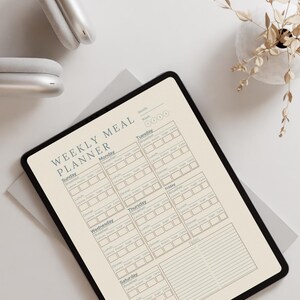 Könnte beinhalten: Ein digitaler Wochenessensplaner auf einem Tablet. Der Planer ist beige mit einem schwarzen Rand und enthält Abschnitte für jeden Wochentag, einschließlich Frühstück, Mittagessen und Abendessen. Der Text "WEEKLY MEAL PLANNER" steht oben. Ein weißer Kopfhörer befindet sich im Hintergrund.